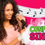 Young Black woman with curly hair smiling while holding a microphone, with music notes and bold text “How to Become a Confident Singer” — visual promoting confidence tips for neurodivergent and sensory-sensitive singers.